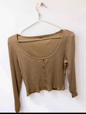 Beige ribbad croppad topp med knappar - Trendig croppad topp i beige ribbad bomull med långa ärmar och knappar framtill. Perfekt för att styla med höga jeans eller kjol. Djup rund halsringning ger en snygg look.