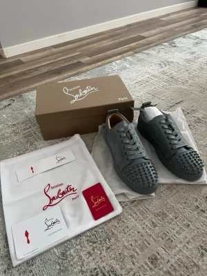 Grå Christian Louboutin sneakers - Säljer ett par gråa Christian Louboutin sneakers i lågt utförande. Skorna är tillverkade i läder och har ikoniska nitar på tån. Klassisk snörning och exklusiv design som sticker ut. Perfekta för dig som vill ha något unikt och trendigt.