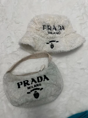 Ljust fluffigt sett med handväska och buckethat - I mycket använt skick, dragkedjan är i sönder!! Supermjuk och fluffig handväska  i ljus färg teddy-material. Väskan har ett broderat svart text -tryck på framsidan. Perfekt accessoar för dig som gillar trendiga och mysiga detaljer.