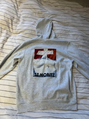 Ljusgrå hoodie med St. Moritz-tryck - Säljer en ljusgrå hoodie. På ryggen finns ett färgstarkt tryck med motiv av snötäckta berg och texten 'ST. MORITZ'. Hoodien har en klassisk känguruficka framtill och är gjord i mjukt sweatshirtmaterial. Perfekt för dig som gillar streetwear och coola prints.