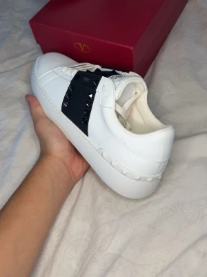 Valentino open/rockstud - Snygga vita sneakers i läder med en bred svart rem över vristen. Skorna har svarta spikar för en look som sticker ut. Perfekta för dig som vill ha en grisch och trendig look.