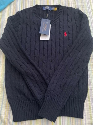 Mörkblå kabelstickad tröja Polo Ralph Lauren - Kabelstickad mörkblå tröja från Polo Ralph Lauren med röd broderad logga på bröstet. Tröjan har v-ringad hals, långa ärmar och ribbade muddar vid ärmslut och nederkant. Perfekt för en klassisk och stilren look.