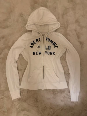 Vit zip up hoodie  Abercrombie fitch - Säljer en vit hoodie med dragkedja från Abercrombie. Tröjan har huva med dragsko, två fickor framtill och mörkblå text med 'Abercrombie 18 New York' broderat på bröstet