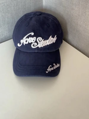 Acne Studios keps - Snygg mörkblå keps från Acne Studios med broderad logga i vitt framtill och på skärmen. Klassisk böjd skärm och justerbar baksida. Tillverkad i mjukt bomullstyg som ger en avslappnad och trendig look.