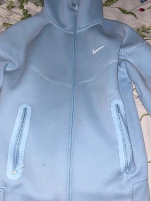Ljusblå NOCTA Nike hoodie med dragkedja - Snygg ljusblå hoodie från NOCTA x Nike med hel dragkedja framtill, två fickor med dragkedja och klassisk huva. Tillverkad i mjukt material och har en vit Nike-logga på bröstet. Perfekt för en sportig och avslappnad stil.