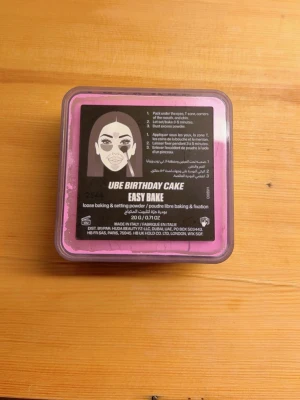Huda Beauty Easy Bake Ube Birthday Cake - Lös setting powder från Huda Beauty i färgen Ube Birthday Cake. Kommer i en fyrkantig svart plastburk med rosa detaljer. Perfekt för baking och fixering av foundation, ger en matt finish och håller sminket på plats hela dagen.