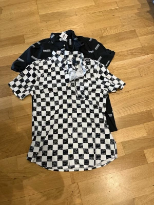 Två kortärmade skjortor från Abercrombie & fitch - Säljer två skjortor i modern stil. Ena skjortan har svartvitt rutigt mönster och den andra är svart med vitt textmönster. Båda har korta ärmar, klassisk krage och knäppning framtill. Perfekta för dig som gillar att sticka ut med grafiska prints.