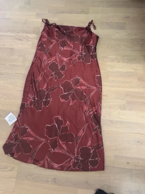 Röd långklänning med blommigt mönster - Snygg röd långklänning med tunna axelband och stora blommor i mörkrött och rosa. Klänningen har en rak passform och är tillverkad i ett glansigt material som ger en elegant känsla. Perfekt för sommarens alla tillfällen.