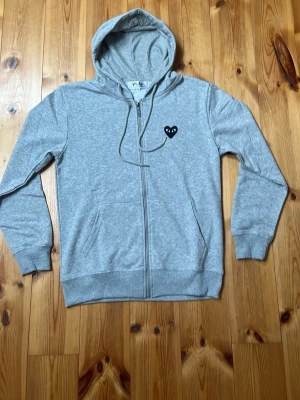Grå hoodie Comme des Garçons Play - Säljer en grå zip-hoodie från Comme des Garçons Play med svart hjärta och ögon på bröstet. Tröjan har huva med dragsko, två fickor fram och ribbade muddar. Perfekt för en chill och stilren look.