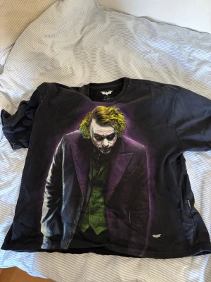 Svart Joker t-shirt från YoungLA - Säljer en svart t-shirt från YoungLA med ett stort tryck av Jokern från The Dark Knight på framsidan. T-shirten har rund hals och korta ärmar. Materialet är mjukt och skönt, perfekt för dig som gillar Batman eller vill ha en unik statement-tee.