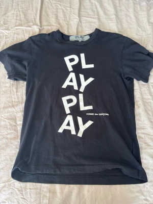 Svart PLAY t-shirt Comme des Garçons - Säljer en svart t-shirt från Comme des Garçons PLAY med stort vitt PLAY-tryck på framsidan. Klassisk rund hals och korta ärmar. Perfekt för dig som gillar streetwear och vill ha något stilrent men ändå med statement.