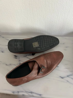 Bruna loafers med spänne - Snygga bruna loafers i läder med dekorativt metallspänne över vristen. Skorna har en klassisk, något spetsig tå och svart sula med diskret mönster undertill. Perfekta för en stilren och avslappnad look.