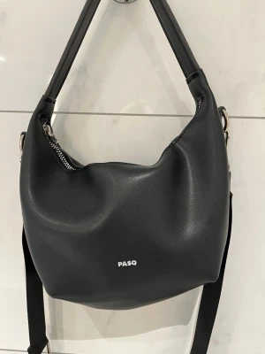 Svart axelväska från PASQ - Snygg svart axelväska från PASQ med minimalistisk design i mycket bra skick. Väskan har en mjuk, rundad form och justerbar axelrem. Tillverkad i slätt konstläder med silverfärgade metalldetaljer och dragkedja upptill. Perfekt för dig som gillar stilrena accessoarer🖤