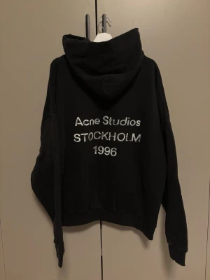Svart hoodie Acne Studios Stockholm - Svart hoodie från Acne Studios med stor huva och tryck i vitt på ryggen där det står 'Acne Studios STOCKHOLM 1996'. Oversized passform och mjukt material, perfekt för en chill och trendig look.
