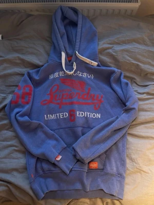 Blå Superdry hoodie med tryck - Snygg blå hoodie från Superdry med röd och vit text på bröstet, japanska tecken och siffran 68 på ärmen. Stor känguruficka framtill och vita dragsnören i huvan. Limited Edition-tryck och detaljer i rött. Perfekt för en avslappnad streetwear-look.