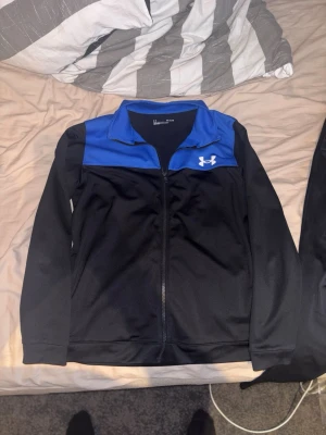 Under Armour svart och blå träningsset - Säljer ett träningsset från Under Armour med en svart och blå zip-jacka och svarta träningsbyxor. Jackan har en blå axelpanel och vit logga på bröstet, byxorna har vit logga på låret. Perfekt för gym eller sport. Materialet är mjukt och stretchigt.
