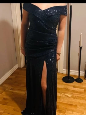 Marinblå glittrig långklänning - Säljer en marinblå långklänning med glittriga paljetter, offshoulder och en snygg slits framtill. Klänningen har en figurnära passform och är perfekt för bal eller fest. Materialet känns lyxigt och skimrar i ljuset. Andvänd en gång!