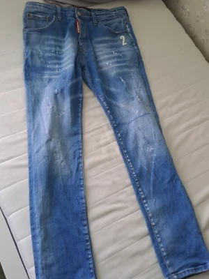 Blå denimshorts med slitningar - Säljer ett par blå denimshorts med slitna detaljer och ljusa tvättade partier. Shortsen har klassisk femficksdesign, bälteshällor och en vit siffra '2' på höger ficka. Perfekta för en avslappnad stil.