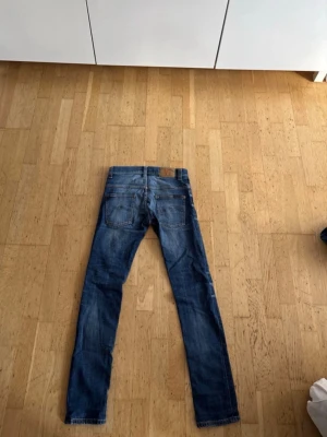 Nudie jeans - Säljer ett par blå slim fit jeans i klassisk denim med snygga slitningar och tvättade detaljer. Jeansen har fem fickor, bälteshällor och en patch bak i midjan. Perfekta för en avslappnad och trendig look.