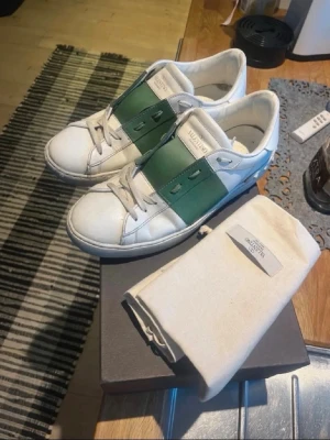 Valentino vita sneakers med grön detalj - Snygga vita sneakers från Valentino med bred grön läderdetalj över snörningen. Klassisk låg modell med rund tå och vita skosnören. Tillverkade i läder och har diskret logga på plösen. Perfekta för dig som vill ha en stilren men ändå unik look. Dustbag och box förekommer tyvärr inte 