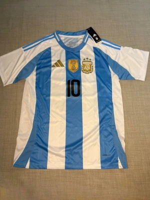 Argentina Messi matchtröja blåvit - Säljer en  Argentina fotbollströja från Adidas med Messi och nummer 10 på ryggen. Tröjan har klassiska blå och vita ränder, korta ärmar och broderade märken på bröstet. Perfekt för dig som älskar fotboll och vill stötta ditt favoritlag!