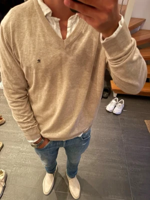 Beige stickad tröja med V-ringning - Säljer en stilren beige stickad tröja med V-ringning och diskret broderad logga på bröstet. Tröjan har långa ärmar och ribbade muddar vid ärmslut och nederkant. Perfekt att bära över en skjorta för en clean look.