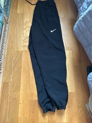 Svarta byxor från Nike med snörning - Säljer ett par svarta shorts från Nike med elastisk midja och snörning. Shortsen har en klassisk passform och är tillverkade i ett lätt och bekvämt material, perfekt för träning eller chill. Nike-loggan syns tydligt på ena benet.
