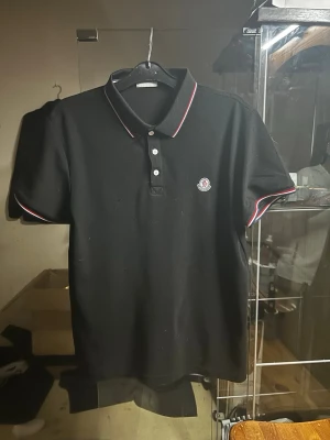 Svart Moncler pikétröja  - Klassisk svart pikétröja från Moncler med vit och röd kant på kragen och ärmsluten. Tröjan har korta ärmar, tre knappar framtill och Moncler-logga på bröstet. Perfekt för en clean och stilren look.