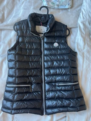 Svart dunväst från Moncler - Snygg svart dunväst från Moncler med hög krage och två dragkedjeförsedda fickor. Västen har quiltad design och Moncler-logga på bröstet. Perfekt för lager-på-lager och håller dig varm med sin lätta dunfyllning.