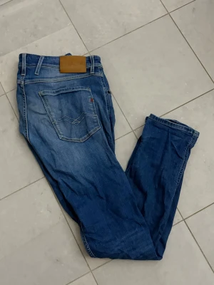 Blå jeans med slitningar från replay - Säljer ett par blå jeans från replay med snygga slitningar och klassisk femficksmodell. Jeansen har normal midja, raka ben och detaljerade sömmar på bakfickorna. Tillverkade i bomull med en skön stretch för extra komfort.