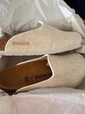 Beige tofflor från Birkenstock - Säljer ett par stilrena beige tofflor från Birkenstock i storlek 38. De har en mjuk ovandel i filtmaterial och klassisk korksula med vit yttersula. Perfekta för att chilla hemma eller på jobbet. Birkenstock-logga broderad på sidan.