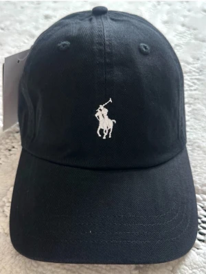 Svart keps från Polo Ralph Lauren - Snygg svart keps från Polo Ralph Lauren med klassisk vit broderad logga framtill och 'POLO' broderat bak. Justerbart spänne i metall baktill för perfekt passform. Tillverkad i bomullstyg som känns både stilrent och bekvämt.