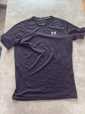 Svart kompressionströja Under Armour - Säljer en svart kompressionströja från Under Armour med HeatGear-teknologi. Tröjan har korta ärmar, rund hals och en vit logga på bröstet. Tillverkad i ett stretchigt funktionsmaterial som andas och passar perfekt till träning.