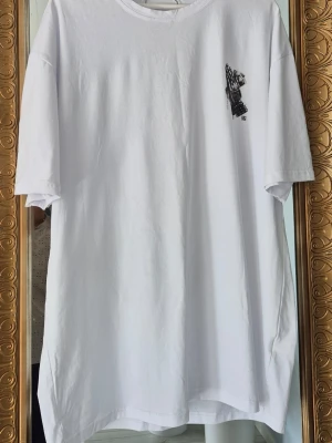 Vit  t-shirt med tryck - Säljer en vit t-shirt med ett stort svart tryck på ryggen som föreställer händer med smycken och sedlar. T-shirten har korta ärmar och är helt ny