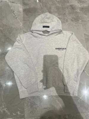 Grå Essentials Fear of God hoodie - Snygg ljusgrå hoodie från Essentials Fear of God med svart logga på bröstet och stor text på ryggen. Klassisk passform med huva och ribbade muddar. Perfekt för en chill och trendig streetwear-look.