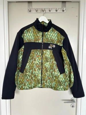 Mountain Hardwear x Stüssy Fleece - Unik fleecejacka i grönt och svart från Mountain Hardwear x Stüssy. Jackan har hel dragkedja, hög krage och två sidofickor. Mönstret är melerat i gröna toner, med logga på bröstet. Perfekt för dig som gillar streetwear och outdoor-vibe.