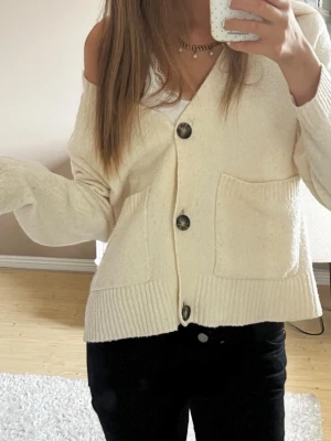 Krämvit kofta från Vero Moda - Mysig krämvit kofta från Vero Moda med stora knappar framtill och två rymliga fickor. Koftan har en avslappnad passform, ribbade muddar och är tillverkad i ett mjukt stickat material. Perfekt för lager på lager och enkel att styla.