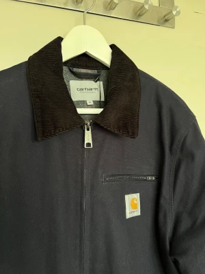 Carhartt Detroit Jacka - Snygg mörkblå Carhartt Detroit jacka med svart manchestertyg på kragen. Jackan har dragkedja framtill, bröstficka med dragkedja och två sidofickor. Tillverkad i slitstarkt tyg och har Carhartt-logga på bröstet. En innerficka.