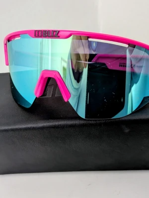 Sportiga rosa solglasögon från Bliz - Säljer ett par sportiga solglasögon från Bliz med rosa bågar och spegelblått glas. De har en modern, svepande form som täcker bra och passar perfekt för utomhusaktiviteter. Riktigt snygga och trendiga för dig som vill sticka ut.