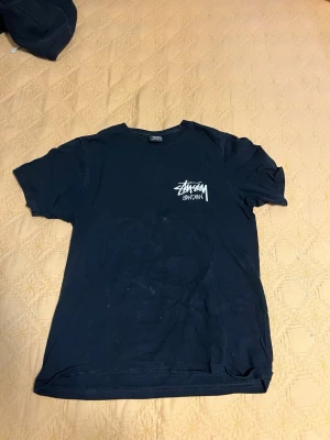 Svart Stüssy t-shirt med tryck - Svart t-shirt från Stüssy med klassiskt logotryck på bröstet. T-shirten har rund hals och korta ärmar, tillverkad i mjuk bomull. Perfekt för en streetwear-look och enkel att matcha med jeans eller shorts.