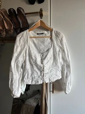 White Embroidered Puff Sleeve Corset Top - This is a white, long-sleeve blouse from Gina Tricot, size M. It features puffed sleeves, a square neckline, and a corset-style lace-up front. The fabric has a subtle floral embroidery pattern, giving it a romantic and vintage-inspired look. Perfect for pairing with high-waisted jeans or skirts for a chic, feminine outfit.
