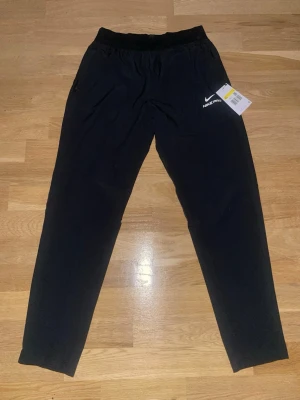 Svarta Nike mjukisbyxor - Svarta mjukisbyxor från Nike med logga på vänster lår och diskret text vid benslutet. Byxorna har en enkel, sportig design med resår i midjan och smal passform. Perfekta för träning eller chill. Snygga och bekväma att bära hela dagen.