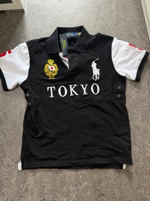 Svart och vit piké med Tokyo-tryck - Säljer en svart och vit pikétröja med krage och knappar. Framtill står det 'TOKYO' i vitt och den har broderad logga samt emblem. Ärmarna är vita med svarta detaljer och röd dekor. Perfekt för dig som gillar sportig stil.