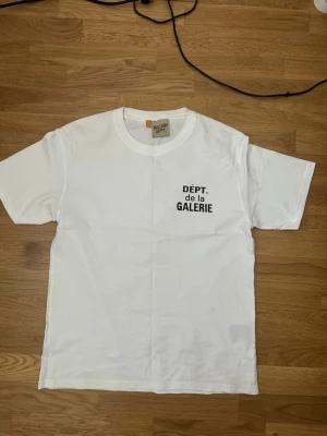 Vit t-shirt från Gallery Dept - Säljer en vit t-shirt från Gallery Dept med svart tryck 'DÉPT. de la GALERIE' både fram och bak. Klassisk rund hals och korta ärmar. Perfekt för dig som gillar streetwear och enkel stil. Den är i storlek M