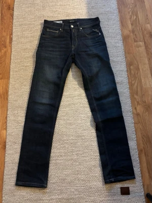 Mörkblå herr jeans från Replay - Snygga mörkblå herr jeans från Replay med klassisk femficksdesign och kontrastsömmar. Jeansen har en läderpatch med Replay-logga bak i midjan och diskreta slitningar framtill. Perfekta för en trendig och avslappnad stil.