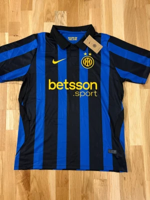 Inter Milan Hemmatröja - Snygg Inter Milan matchtröja från Nike i blått och svart med ränder. Tröjan har korta ärmar, krage och gula detaljer som klubbmärke, Nike-logga och sponsortryck. Tillverkad i lätt och ventilerande material, perfekt för fotboll eller träning.