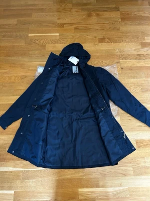 Mörkblå parkas med huva - Stilren mörkblå parkas med huva och tryckknappar framtill. Jackan har fyra fickor med lock och knappstängning samt dragsko i midjan för justerbar passform. Tillverkad i ett slätt, vattenavvisande material. Perfekt för kyliga dagar.