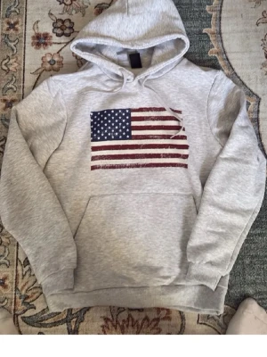 Grå hoodie med amerikansk flagga - Snygg grå hoodie med stor amerikansk flagga tryckt på bröstet. ! Pris kan diskuteras😊helt oanvänd