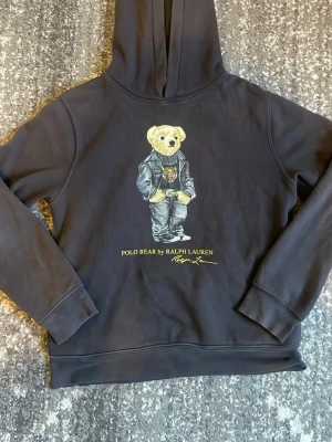Svart hoodie Polo Bear Ralph Lauren - Svart hoodie från Ralph Lauren med den ikoniska Polo Bear-trycket på bröstet. Björnen har på sig en cool outfit med skinnjacka och tigertröja. Hoodien har huva och ribbade muddar. Perfekt för dig som gillar streetwear och vill sticka ut. Storlek 14-16 som ör som xs. Mycket bra skick