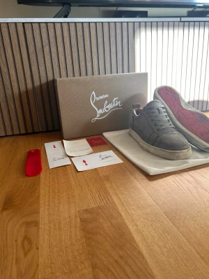 Gråa sneakers från Christian Louboutin - Säljer ett par gråa sneakers från Christian Louboutin med klassisk röd sula och lågt skaft. Skorna är i mocka och har vita sulor. Medföljer originalkartong, skohorn och äkthetsintyg. Perfekta för dig som vill ha något exklusivt och stilrent.